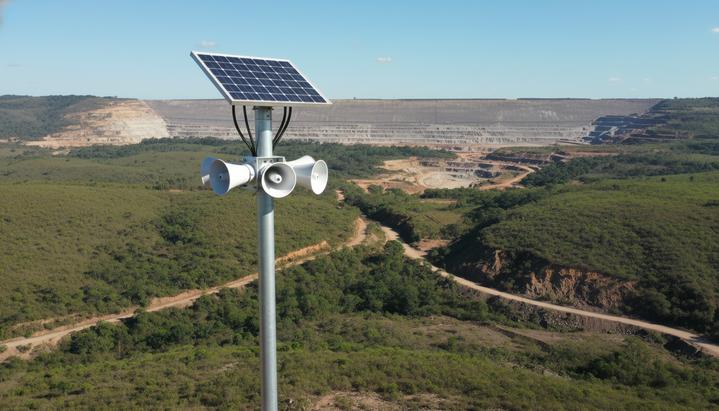 Mineração Morro do Ipê внедрила систему HÖRMANN для предупреждения о прорыве дамбы Современная сирена оповещения с солнечной панелью на мачте на фоне холмистого ландшафта и дамбы хвостохранилища.